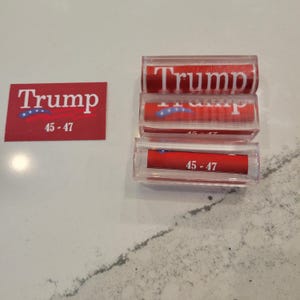 May include: Three red and white plastic tubes with the text "Trump 45-47" printed on them. A red and white sticker with the text "Trump 45-47" is also shown.