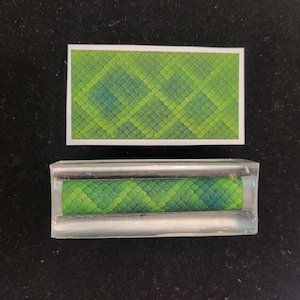 May include: A rectangular green and blue patterned design, resembling snake scales, is displayed on a white background. Below, the same pattern is encased in a clear, rectangular block. The items are set against a dark surface.