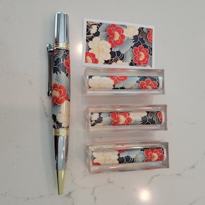 May include: A silver pen with a black and blue floral design. The pen is displayed with three clear acrylic boxes, each containing a piece of fabric with a similar floral design.
