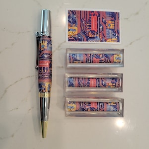 May include: A silver pen with a blue, red and yellow circuit board design. The pen is displayed with three clear acrylic tubes, each containing a similar circuit board design.