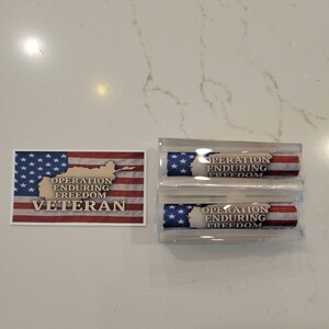 May include: An American flag design with the words "Operation Enduring Freedom Veteran" is featured on a sticker and two cylindrical objects. The objects are in a clear plastic case. The flag design includes stars and stripes.
