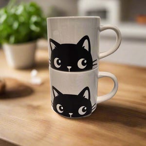 May include: Two white ceramic mugs with black cat faces peeking over the rim. The mugs are stacked on top of each other.