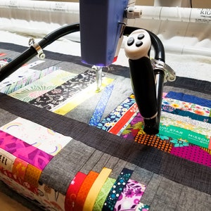 May include: A close-up of a longarm quilting machine in use, stitching a colourful patchwork quilt. The machine has two handles and a needle that is moving across the fabric. The quilt is made of many different fabrics, including floral prints, stripes, and solids. The quilt is being stitched in a straight line.