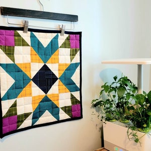 May include: A colorful quilt block with a star pattern in shades of teal, yellow, white, and black. The quilt block is framed with a black border and has pink squares in the corners.
