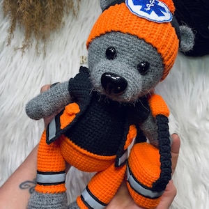 Może przedstawiać: Ręcznie robiony szydełkowy miś ubrany jako ratownik medyczny. Miś jest szary z pomarańczową czapką i spodniami, czarną kamizelką i odblaskowymi paskami. Czapka ma białą naszywkę z niebieskim symbolem medycznym.