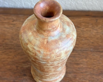May include: A small ceramic vase with a narrow neck and a rounded body, featuring a mottled glaze in shades of tan, beige, and light brown with a textured, earthy appearance and a smooth, circular opening at the top.