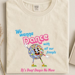 May include: A cream-colored t-shirt featuring a graphic of a disco ball character with arms and legs. The text reads "We wanna Dance with all our friends" in blue and pink, and "Dj's Dont Dance No More" in pink at the bottom. The shirt has a Comfort Colors label.