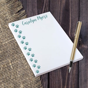 May include: White notepad with a teal paw print border and the name "Carolyn Myers" printed at the top. A gold pen is lying next to the notepad on a brown wooden surface.