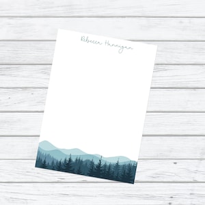 May include: A white notepad with a blue mountain and forest silhouette at the bottom. The name "Rebecca Hannigan" is written in blue cursive at the top of the notepad.