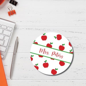 May include: White round coaster with a green and white stripe and the text "Mrs. Peters" in red cursive. The coaster is decorated with a pattern of red apples with green leaves on a white background.