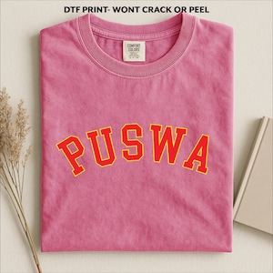 May include: A pink t-shirt with the word "PUSWA" in red block letters with a yellow outline. The shirt is folded and has a Comfort Colors tag. The text "DTF PRINT- WONT CRACK OR PEEL" is at the top.