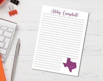 May include: A white lined notepad with the text "Abby Campbell" at the top and a purple outline of the state of Texas at the bottom. The notepad is on a white wooden surface.