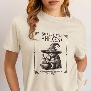 May include: A cream-colored short-sleeve t-shirt featuring a black graphic print of a cat wearing a witch hat and stirring a cauldron. The text reads: SMALL BATCH HEXES, HANDCRAFTED MISCHIEF, EST. 1692. The design is enclosed in a rectangular border.