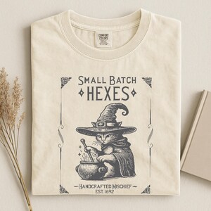 May include: A cream-colored t-shirt featuring a dark gray graphic of a cat wearing a witch hat and cloak, stirring a cauldron. The text reads "SMALL BATCH HEXES" and "HANDCRAFTED MISCHIEF EST. 1692" with decorative border elements.