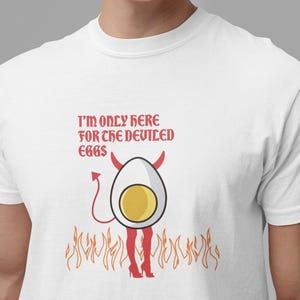 May include: White t-shirt with a humorous graphic. The design features a deviled egg character with horns, a tail, and red high heels, standing in flames. The text reads, "I'M ONLY HERE FOR THE DEVILED EGGS."