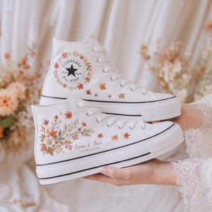Op de afbeelding: Een paar witte hoge canvas sneakers met geborduurde herfstbladeren en bloemmotieven. De zijkant van één schoen toont de tekst Emma & Jack 10.05.2026 in een cursief lettertype, terwijl de andere het ronde Converse All Star logo bevat.