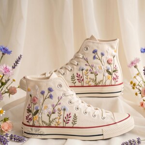 Puede incluir: Un par de zapatillas altas de lona color hueso con intrincados bordados florales. El diseño muestra flores silvestres coloridas en tonos rosa, azul, amarillo y verde. Los zapatos tienen cordones blancos, ojales plateados y una raya roja y negra.