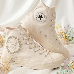 May include: A pair of off-white high-top canvas sneakers featuring a circular embroidered floral wreath with the letters E & J and the date 07.07.26. The shoes have matching laces, silver eyelets, and a circular Converse All Star logo patch.