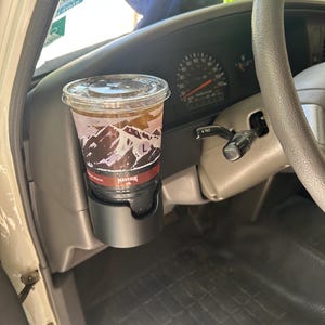 May include: A black plastic cup holder mounted to the dashboard of a vehicle, containing a clear plastic disposable cup filled with a dark iced beverage. The cup features a mountain graphic and the text Maverik printed on the side of the cup.