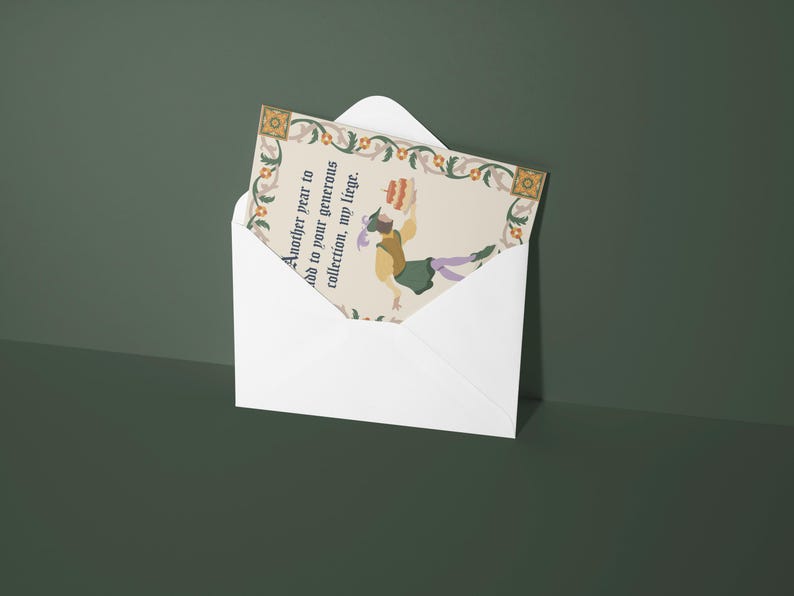May include: A white envelope with a colorful card inside. The card has a floral border and a picture of a person holding a cake. The text on the card reads "Another year to add to your generous collection, my liege."