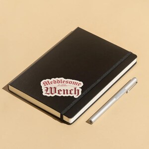 Puede incluir: Un cuaderno negro con una pegatina blanca que dice "Meddlesome little Wench" en letras rojas. Un bol&iacute;grafo plateado est&aacute; al lado del cuaderno.
