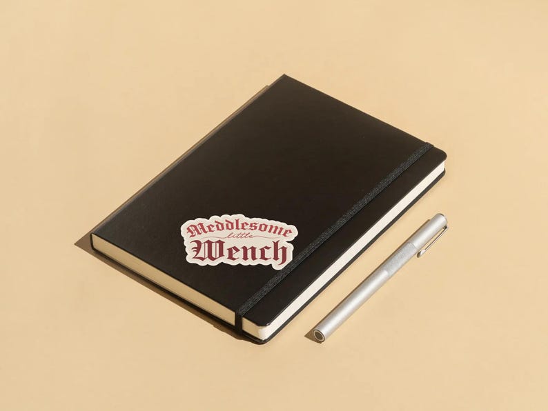 Puede incluir: Un cuaderno negro con una pegatina blanca que dice "Meddlesome little Wench" en letras rojas. Un bol&iacute;grafo plateado est&aacute; al lado del cuaderno.