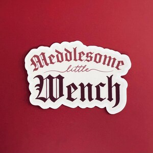 Puede incluir: Pegatina blanca con texto negro que dice "Meddlesome little Wench" en una fuente g&oacute;tica.