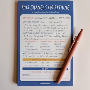 May include: A blue notepad with the text "THIS CHANGES EVERYTHING" and handwritten notes, along with a pink pen. The notepad is designed as a practical guide for big ideas, with sections for idea/obsession, time commitment, and financial impact.