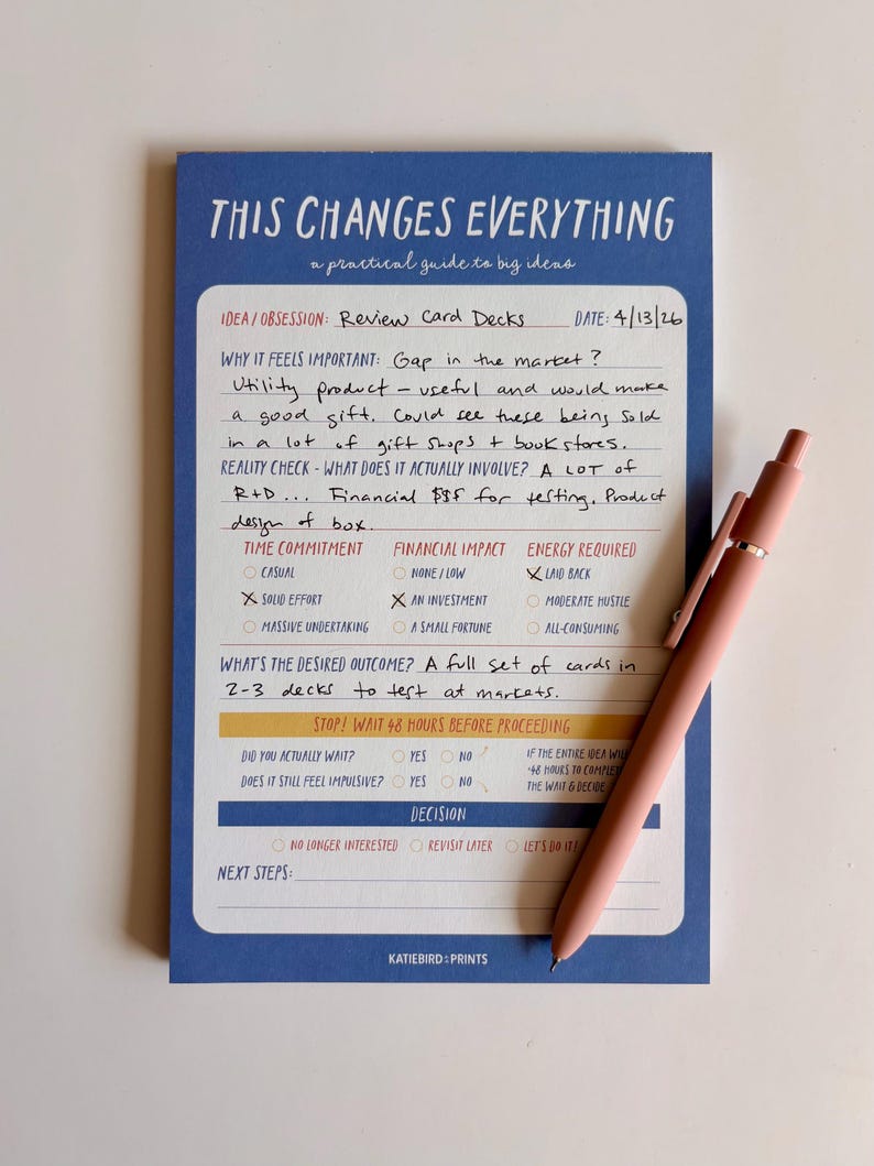 May include: A blue notepad with the text "THIS CHANGES EVERYTHING" and handwritten notes, along with a pink pen. The notepad is designed as a practical guide for big ideas, with sections for idea/obsession, time commitment, and financial impact.