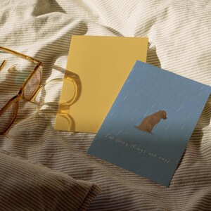 May include: A pair of brown-framed eyeglasses, a yellow card, and a blue card with a dog illustration and the text "I'm sorry things are rough" rest on a striped, cream-colored bedspread.