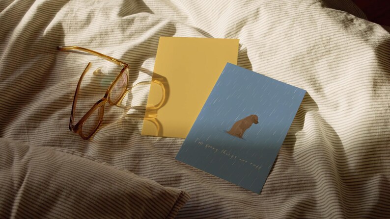 May include: A pair of brown-framed eyeglasses, a yellow card, and a blue card with a dog illustration and the text "I'm sorry things are rough" rest on a striped, cream-colored bedspread.