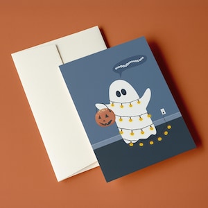 May include: A Halloween-themed greeting card featuring a cartoon ghost holding a jack-o'-lantern and wrapped in string lights. The card is paired with a cream-colored envelope. The background is a muted blue with a brown surface.