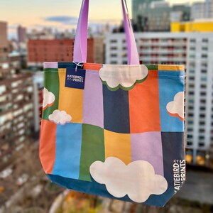 Puede incluir: Una bolsa de tela colorida con un patrón geométrico de rayas y nubes. La bolsa tiene un asa morada y una etiqueta azul con el texto "KATIEBIRD PRINTS".
