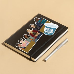 May include: A black notebook with a sticker of a group of people sitting on a bench. The sticker is in a cartoon style and features a variety of skin tones and hairstyles. There is also a sticker of a blue yogurt container with the text "Breeder's Fat Free Yogurt" on it. A silver pen is lying next to the notebook.