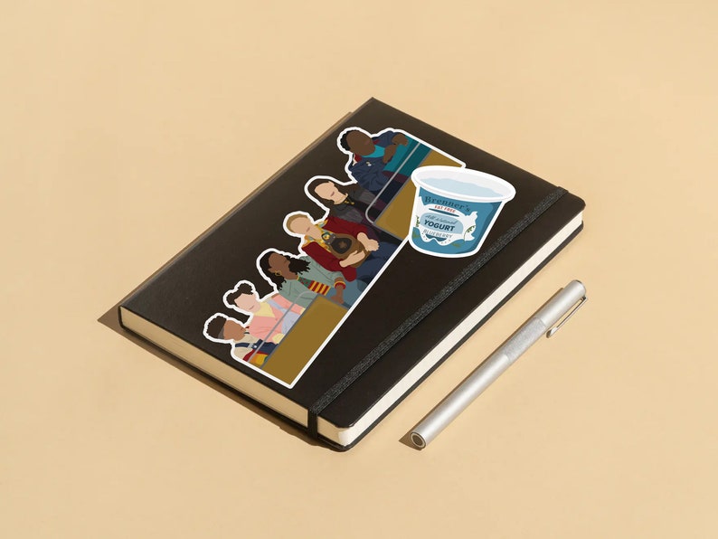 May include: A black notebook with a sticker of a group of people sitting on a bench. The sticker is in a cartoon style and features a variety of skin tones and hairstyles. There is also a sticker of a blue yogurt container with the text "Breeder's Fat Free Yogurt" on it. A silver pen is lying next to the notebook.