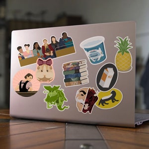 May include: A laptop computer with various stickers on the lid. The stickers include a group of people, a hamster, a monster, a stack of books, a carton of yogurt, a carton of milk, a pineapple, and a creature with a human face and a demon face.