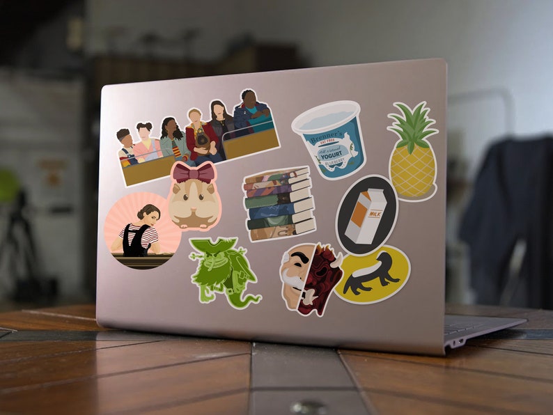 May include: A laptop computer with various stickers on the lid. The stickers include a group of people, a hamster, a monster, a stack of books, a carton of yogurt, a carton of milk, a pineapple, and a creature with a human face and a demon face.