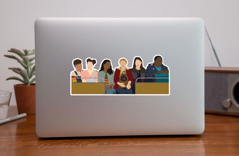 May include: A group of six people, three women and three men, are sitting on a bench. They are all wearing different colored clothing. The people are looking at the camera. The bench is brown and has a gold trim.