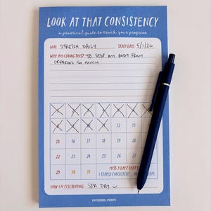 May include: A blue notepad with the text "LOOK AT THAT CONSISTENCY" and a calendar grid. The notepad includes prompts for goals and a start date. A dark blue pen rests on the notepad, which is designed to track progress.
