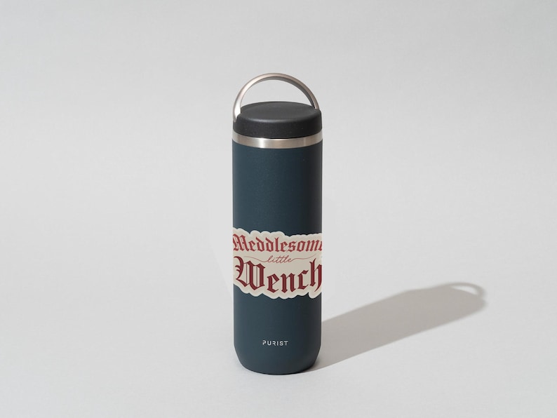 Puede incluir: Una botella de agua de acero inoxidable azul oscuro con tapa negra y asa plateada. La botella tiene una pegatina que dice "Meddlesome little Wench" en una fuente g&oacute;tica roja.