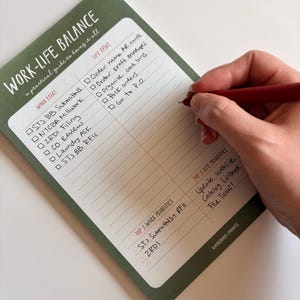 May include: A green and white notepad with the text "WORK-LIFE BALANCE" and a list of tasks. A hand holding a red pen is writing on the notepad. The notepad includes sections for work and life tasks.