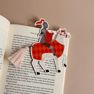 May include: A bookmark featuring a knight in silver armor riding a white horse with red and orange diamond-patterned accents. The bookmark has a pink tassel and is placed on an open book.