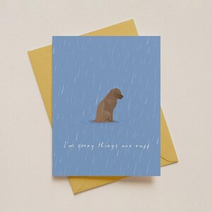 May include: A greeting card with a blue background and a brown dog illustration in the rain. The text "I'm sorry things are ruff" is written in white. The card is on top of a yellow envelope.