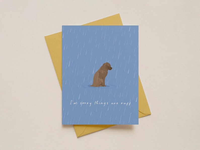 May include: A greeting card with a blue background and a brown dog illustration in the rain. The text "I'm sorry things are ruff" is written in white. The card is on top of a yellow envelope.