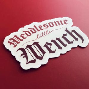 Puede incluir: Pegatina blanca con letras negras y rojas que dicen "Meddlesome little Wench".