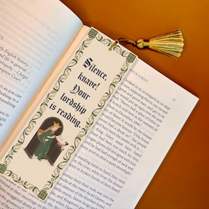 May include: A decorative bookmark with a gold tassel, placed in an open book. The bookmark features the text "Silence, knave! Your lordship is reading." and an illustration of a person in a green robe.