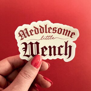 Puede incluir: Una pegatina blanca con el texto "Meddlesome little Wench" en fuente g&oacute;tica roja.