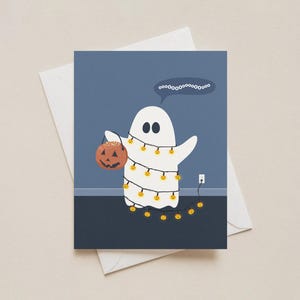 May include: A Halloween-themed greeting card featuring a white ghost wrapped in string lights and holding a jack-o'-lantern. The ghost has a speech bubble with the word "oooooooooooooooo". The card is on a white envelope.