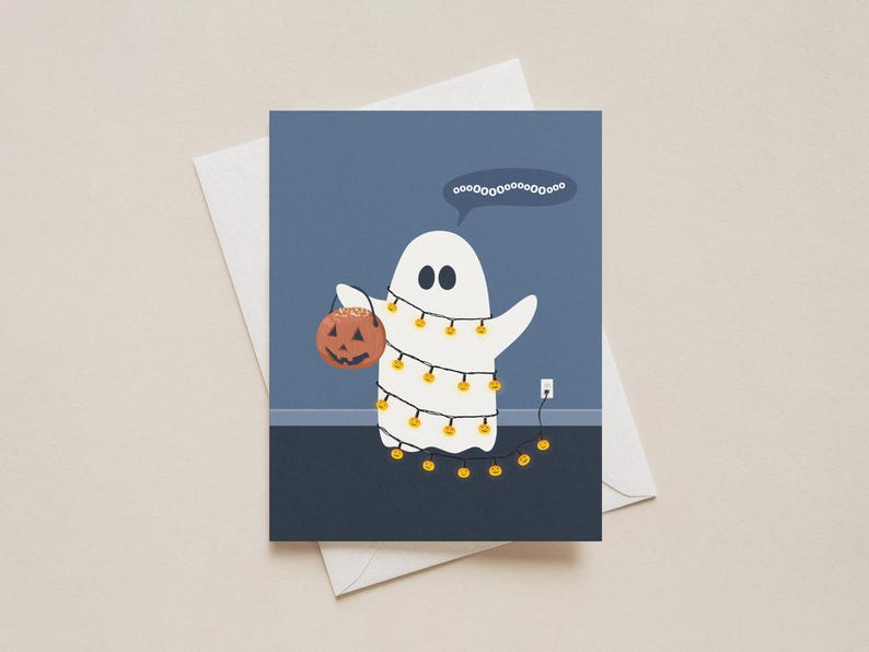 May include: A Halloween-themed greeting card featuring a white ghost wrapped in string lights and holding a jack-o'-lantern. The ghost has a speech bubble with the word "oooooooooooooooo". The card is on a white envelope.