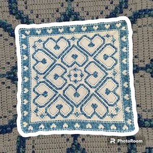 May include: Square crochet piece with a white center and blue heart-shaped patterns. The border is blue with small heart shapes. The background is a gray crocheted surface.